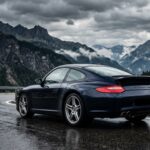 Porsche 997 Carrera 4S bleu foncé sur route de montagne humide, vue de trois-quarts arrière